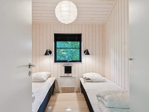 Ein Zimmer mit zwei Betten, einem Fenster mit Jalousien, einer Deckenleuchte und einem an der Wand befestigten Fernseher.