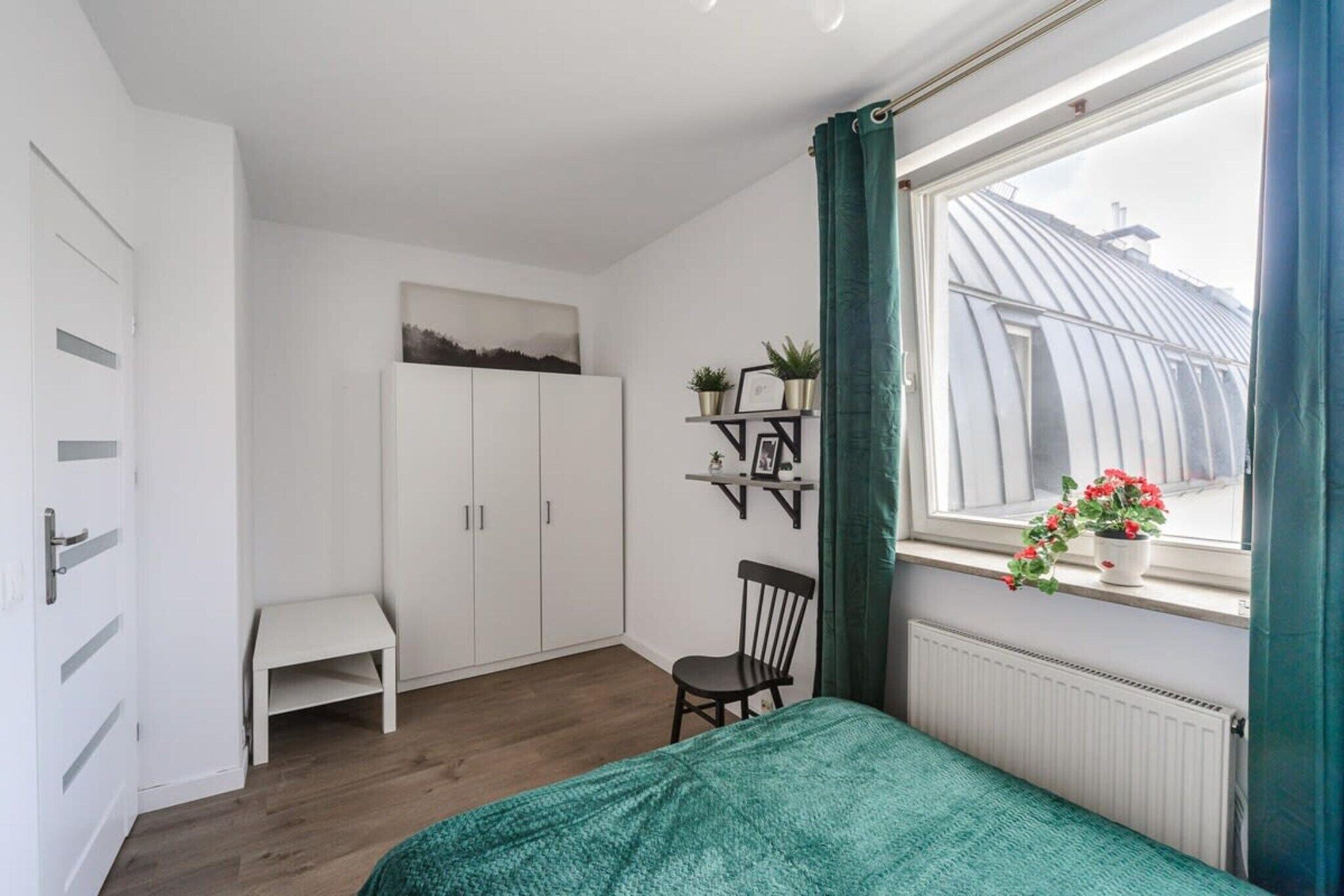 A bedroom with a bed, a chair, a wardrobe, a shelf with plants, and a window with curtains.