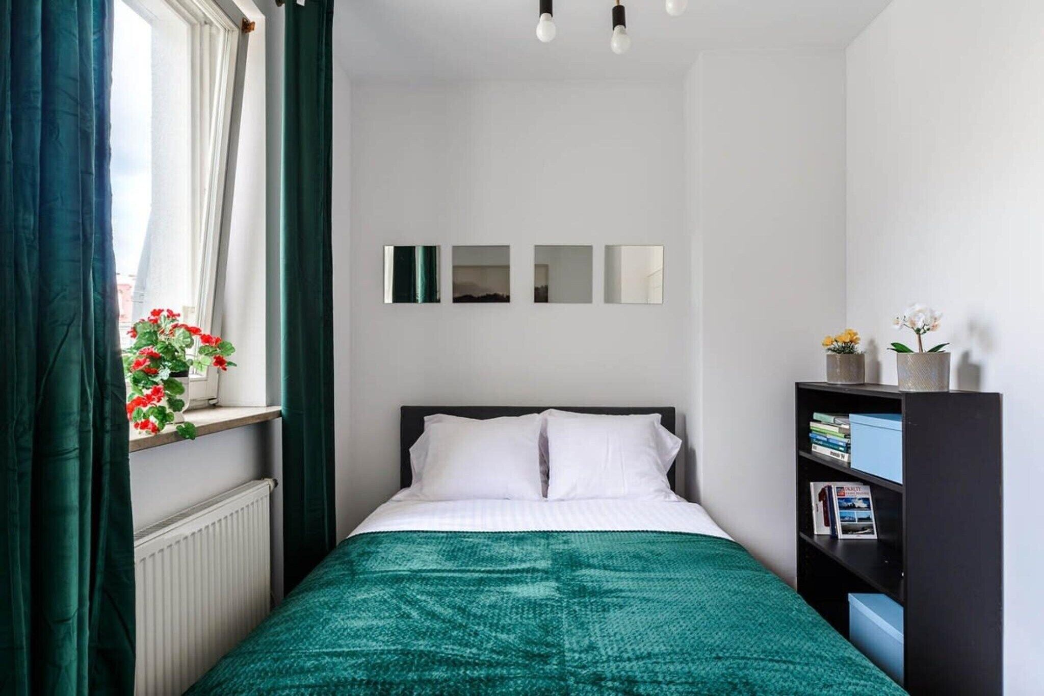 Ein Schlafzimmer mit Bett, einem Vorhangfenster, einem Heizkörper, einem Bücherregal und einem Spiegel an der Wand.