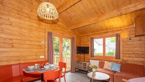A wooden cabin interior with a dining table, chairs, a sofa, a TV, and a window.
