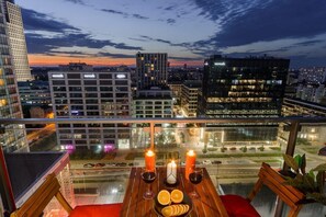 Eine Dachterrasse mit einem Tisch für zwei Personen, Kerzen und Blick auf die städtische Silhouette im Abendlicht.