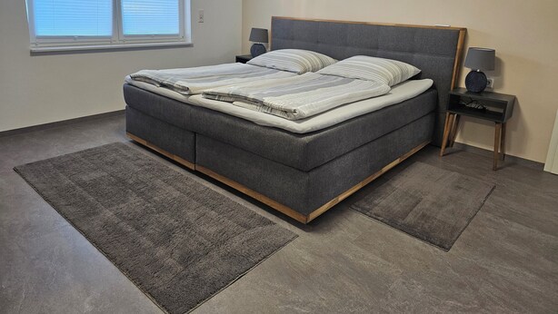 A modern bedroom with a grey upholstered bed, a bedside table with a lamp, and a window with blinds.