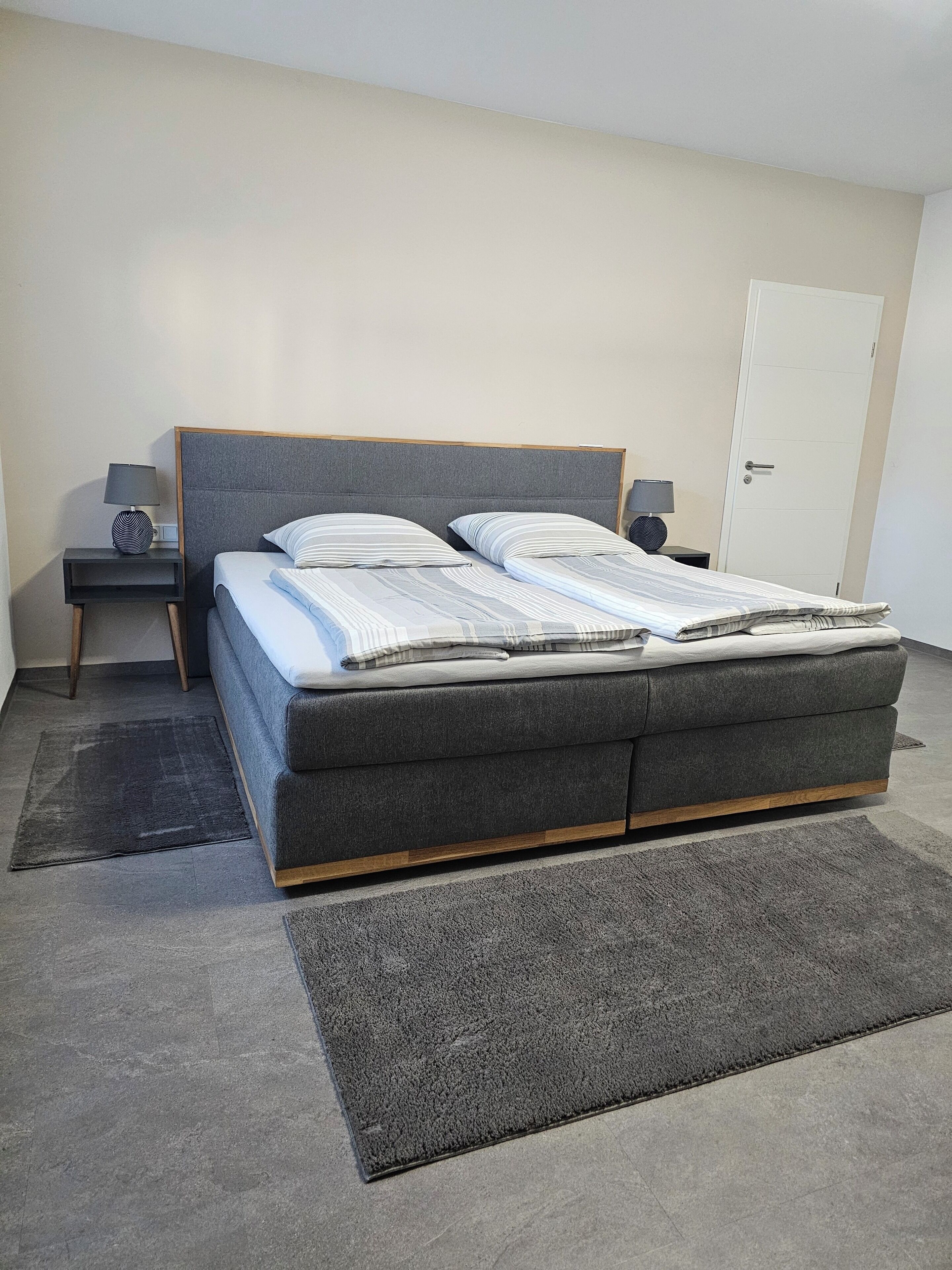 A modern bedroom with a large bed, two bedside tables with lamps, a door, and a carpeted floor.