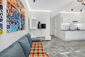 A modern living room with a sofa, a flat-screen TV mounted on the wall, and a kitchen area with white cabinets and a countertop. - Home near Piotrkowska Street (Łódź)