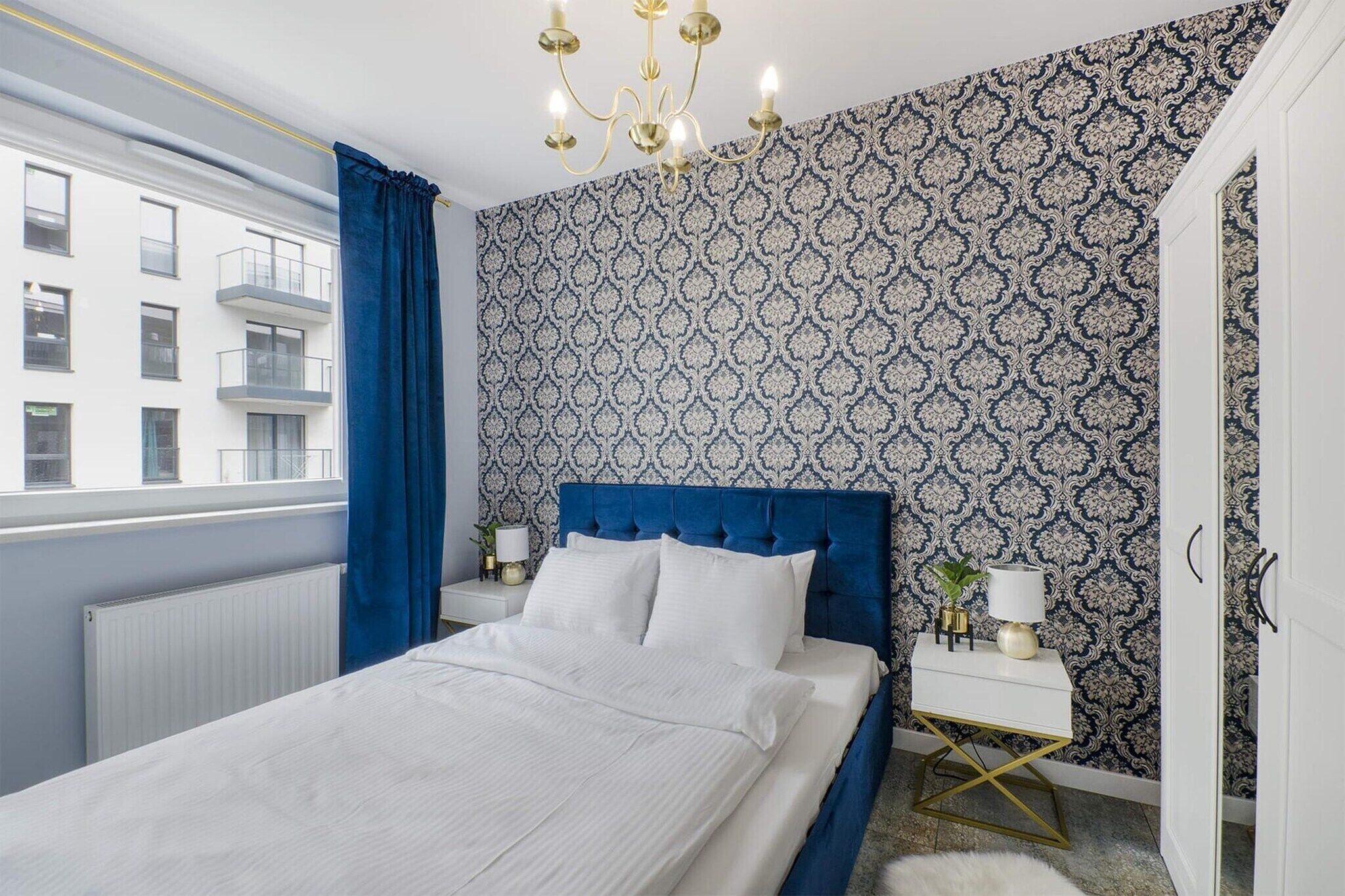 Un dormitorio con cama, mesita de noche, lámpara, ventana con cortinas y un papel tapiz estampado.