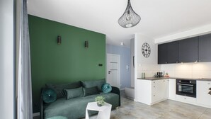 A modern living room with a green sofa, a white side table, and a kitchen area with white cabinets and a black oven. - House by Plage de Melon with Garden Retreat (Łódź)