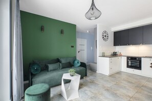 A modern living room with a green sofa, a white side table, and a kitchen area with white cabinets and a black oven. - House by Plage de Melon with Garden Retreat (Łódź)