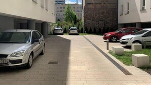 Ein Parkplatz mit parkenden Autos entlang eines befestigten Weges, umgeben von Gebäuden mit Fenstern und einer Ziegelmauer.