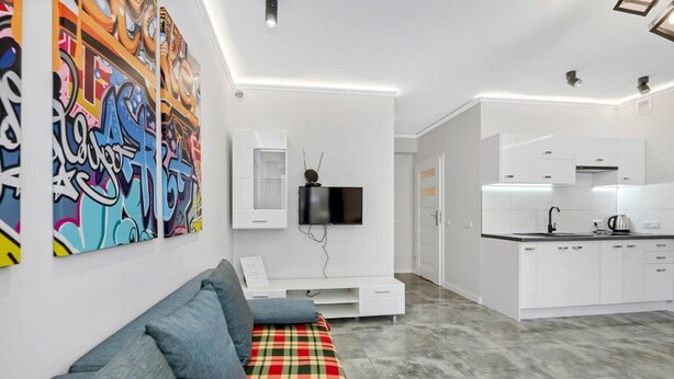 Ein modernes Wohnzimmer mit einer Couch, einem an der Wand befestigten Flachbildfernseher und einer Küchenzeile mit weißen Schränken und einer Arbeitsplatte.