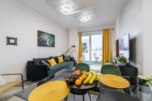 Ein modernes Wohnzimmer mit einer Couch, einem Esstisch mit Früchten und einem großen Fenster mit Vorhängen.