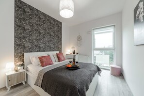 Ett modernt sovrum med en stor säng, ett sängbord, ett litet bord, ett fönster och en vägg med ett lövmönster.