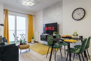 Ein modernes Wohnzimmer mit einem Flachbildfernseher, grünen Sesseln, einem Esstisch und einem gelben Vorhang.