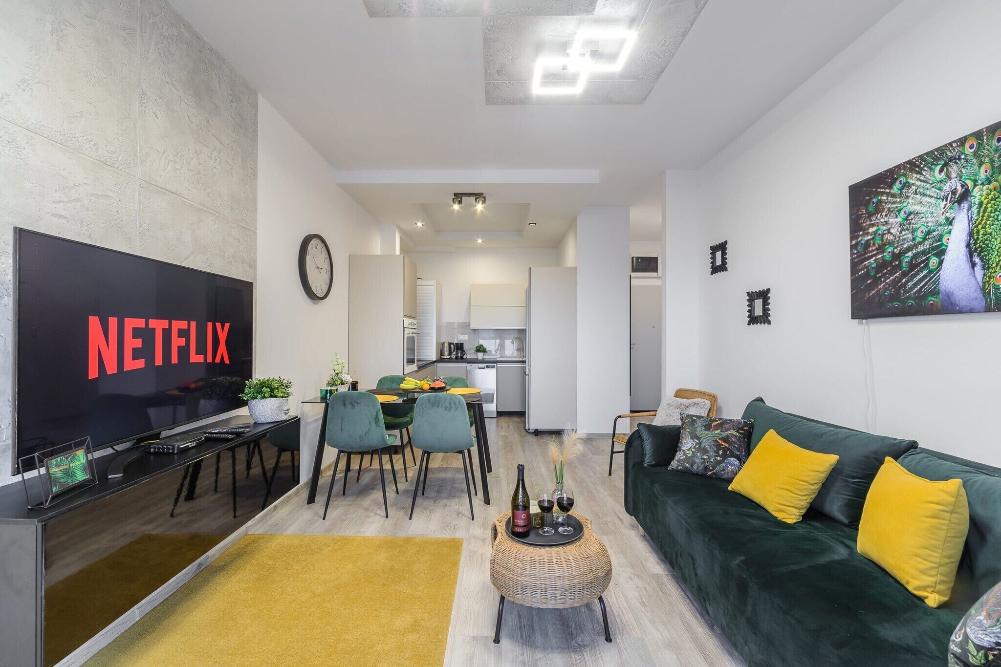 A modern living room with a flat-screen TV, a dining area, and a kitchen in the background.