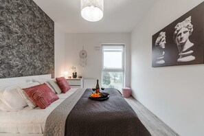 A bedroom with a bed, a bedside table with a lamp, a dreamcatcher, and a window with a view. - City Haven near Atlas Arena (Łódź)