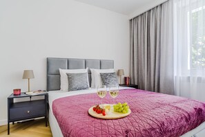 A neatly made bed with a pink quilt, two bedside tables with lamps, a nightstand with a lamp, and a window with curtains. - Charming Apartment near the beach (Warszawa 2)