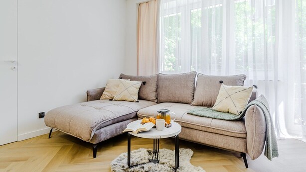 Ein modernes Wohnzimmer mit einem Ecksofa, einem kleinen runden Couchtisch und Blick auf Grün hinter transparenten Vorhängen.
