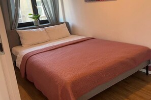 Ein Bett mit einer rosa Decke, ein Fenster mit Vorhängen und ein Topfpflanze am Fensterbrett.