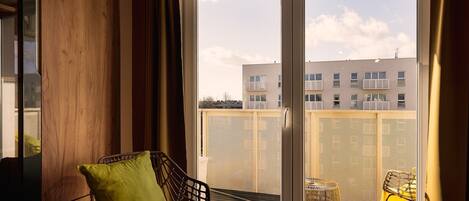Ein Balkon mit einem Korbstuhl, einem kleinen Tisch, einem Metallgeländer und einem Gebäude im Hintergrund.