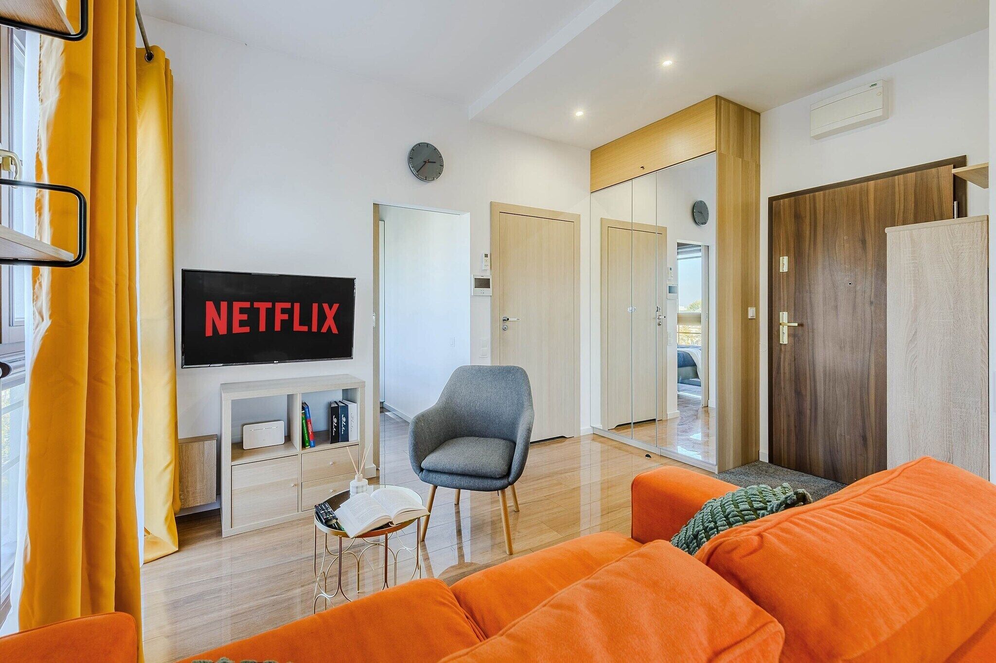 A modern living room with a television displaying 'NETFLIX,' a grey armchair, an orange sofa, and wooden doors leading to other rooms.