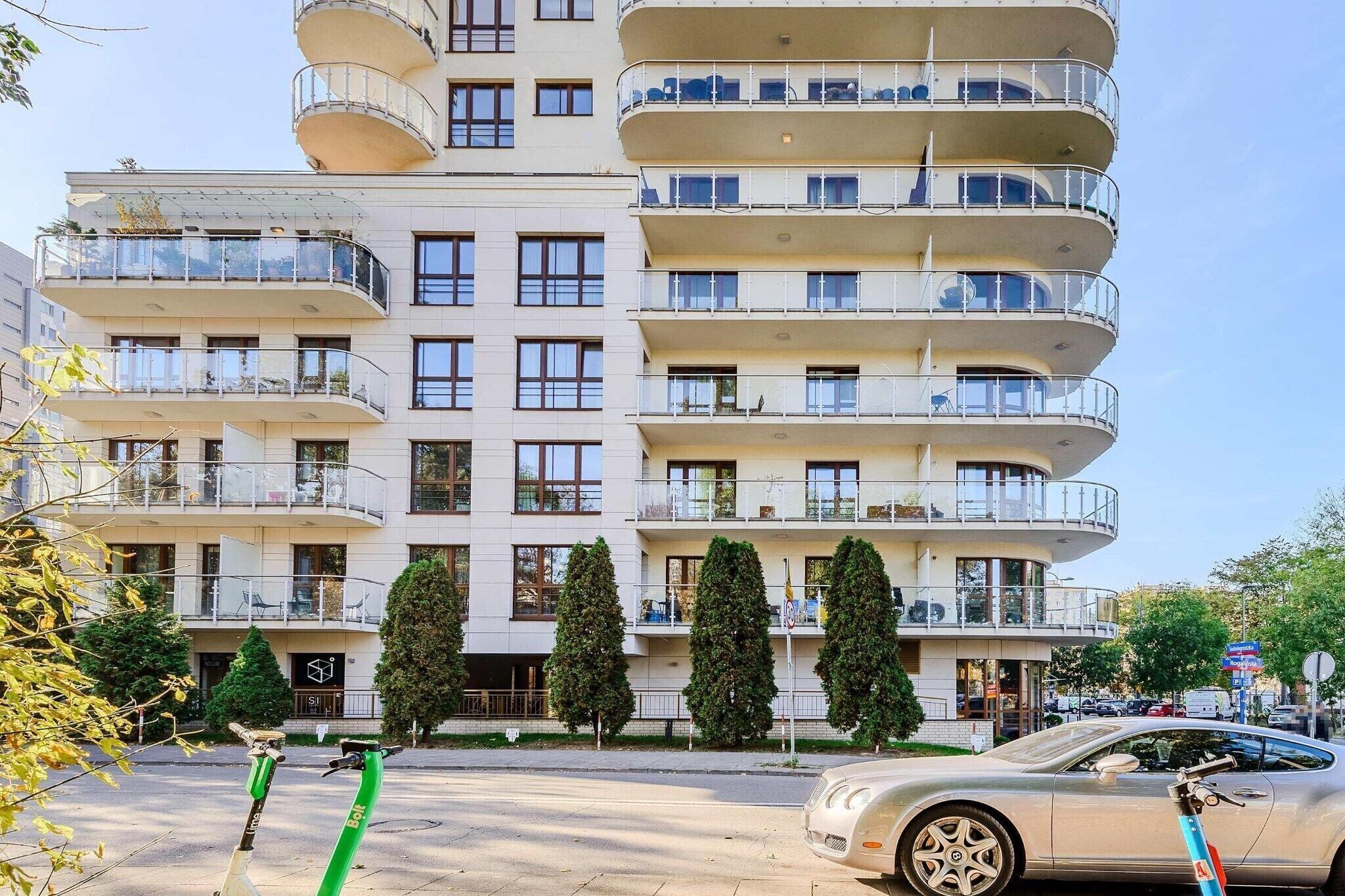 Un edifici d'apartaments de diversos pisos amb balcons, una zona d'aparcament i vista al carrer.