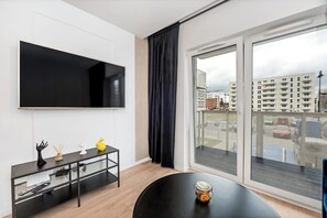 Ein modernes Wohnzimmer mit Flachbildfernseher, einem schwarzen TV-Schrank, einem runden Tisch mit Kerze und einem großen Fenster mit Blick auf Gebäude.