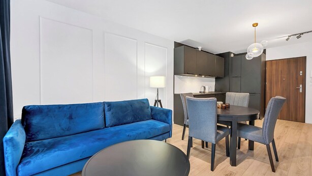 A modern living room with a blue sofa, a dining table with chairs, and a kitchen area. - Elegant Apartment near Vitsula River (Łódź)