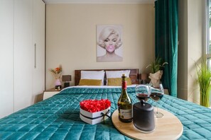 Ein ordentlich bezogenes Bett mit einer petrolfarbenen Steudecke, eine Schachtel roter Rosen, eine Weinflasche und zwei Weingläser auf einem Nachttisch.