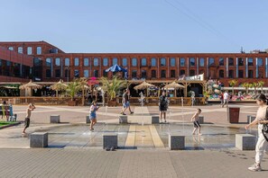 Ein öffentlicher Platz mit einem Wasserspiel, Sitzgelegenheiten und einem Gebäude mit großen Fenstern im Hintergrund.