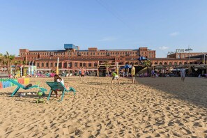 Ein sandiger Strand mit einem großen Backsteinhaus im Hintergrund, einem Volleyballnetz und Liegestühlen.