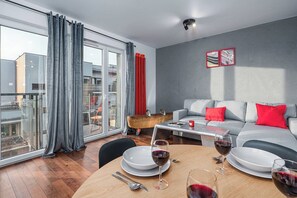 A modern living room with a grey sofa, red cushions, a dining table set with plates and wine glasses, and a balcony with a view of buildings. - Warsaw Flat near Praga Park (Warszawa 2)