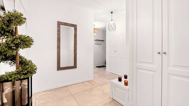 Un couloir aux murs blancs, avec un miroir, une plante en pot et une porte dotée d’une armoire.