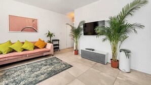 Un salon moderne avec un canapé rose, des coussins verts et jaunes, un téléviseur fixé au mur et des plantes en pot.
