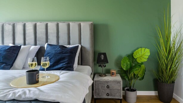 Quarto com cama, mesas de cabeceira, luminária, planta em vaso e bandeja com copos e caneca.