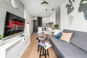 Uma sala de estar moderna com televisão de tela plana, um sofá cinza, uma mesa de centro com revistas e uma cozinha pequena ao fundo.