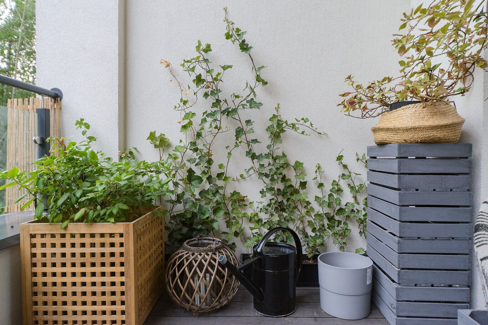 A balcony with a wooden planter box, a black watering can, a grey pot, and a woven basket on a grey wooden structure.