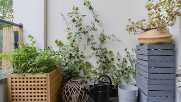 A balcony with a wooden planter box, a black watering can, a grey pot, and a woven basket on a grey wooden structure.
