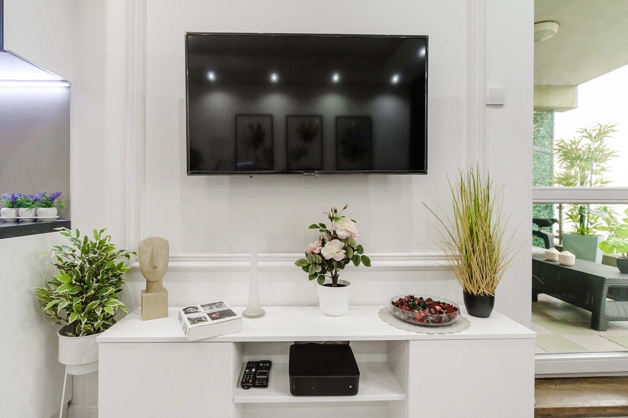Uma sala de estar moderna com uma TV de tela plana na parede, uma mesinha console branca com objetos decorativos e vista para o exterior por uma grande janela.