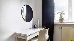 Escritorio blanco con una silla, un espejo redondo, un reloj y una ventana que da a edificios y una planta.
