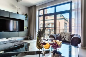 Ein modernes Wohnzimmer mit Fernseher, einer Couch, einem Couchtisch mit Früchten und Getränken sowie einem großen Fenster mit Blick auf die Stadt.
