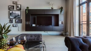 A modern living room with a flat-screen TV mounted on a dark wall, a glass coffee table with fruits and wine glasses, a black leather sofa, and a large window with curtains. - Loft Flat in Historical Area (Łódź)