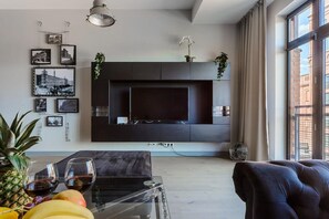 Ein modernes Wohnzimmer mit einem Flachbildfernseher an einer dunklen Wand, einem Glastisch mit Früchten und Weingläsern, einem schwarzen Ledersofa und einem großen Fenster mit Vorhängen.