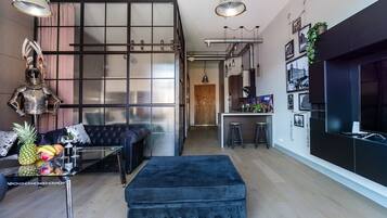 A modern living room with a black sofa, a glass coffee table with fruits, and a blue ottoman.