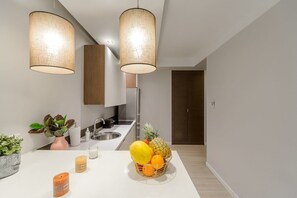 A modern kitchen with a white countertop, two hanging pendant lights, a stainless steel sink, and a fruit bowl with pineapple, mango, and orange. - City Apartment with Balcony (Warszawa)