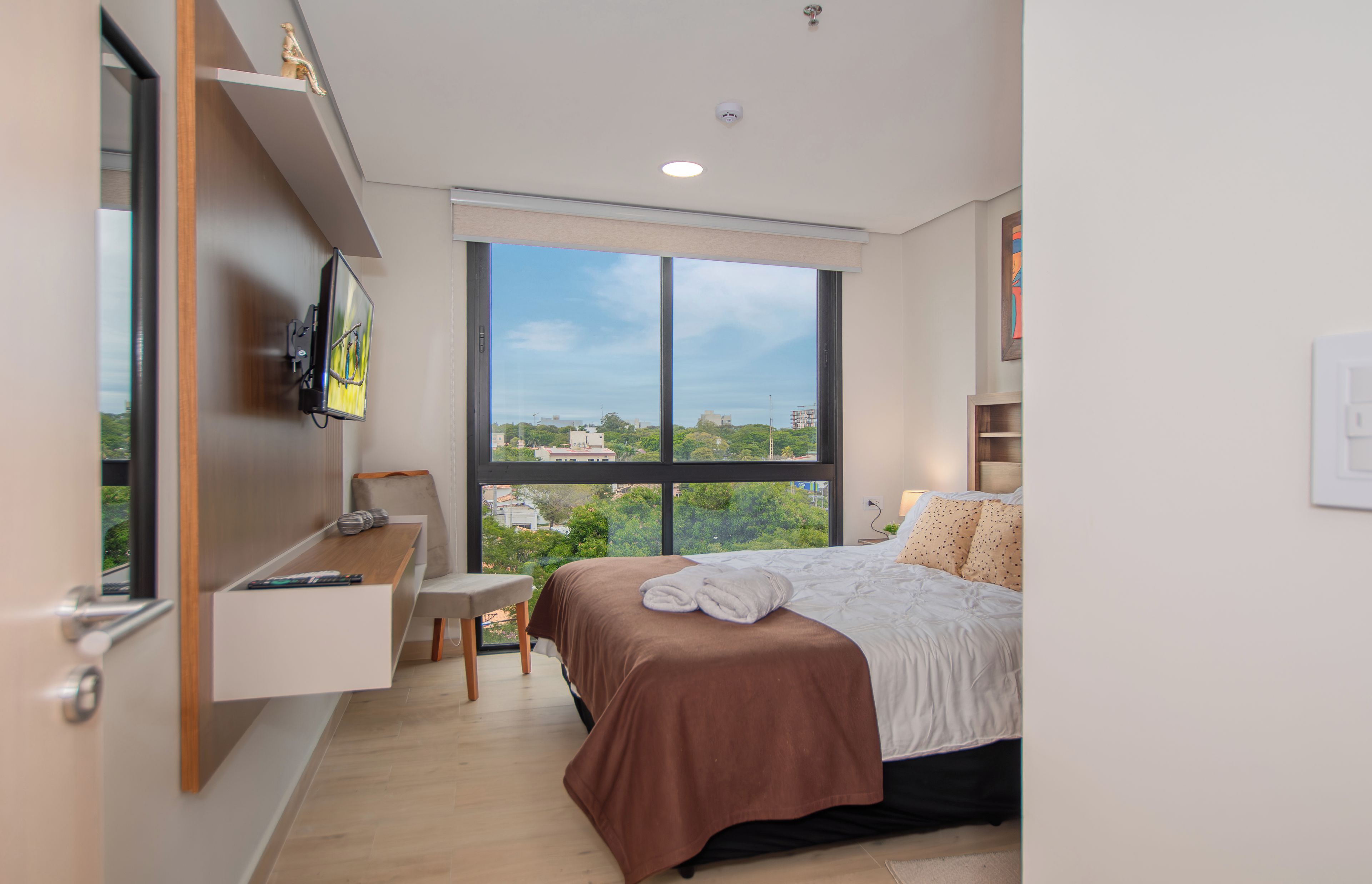 A modern bedroom with a large window, a bed, a desk, and a television.