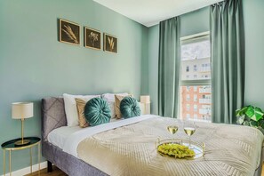 Ein Schlafzimmer mit einem Bett, Kissen, einem Nachttisch mit Lampe, einem Fenster mit Vorhängen und Blick auf Gebäude.