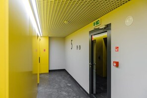 Ein Korridor mit gelber Wand, einer Tür mit einem Notausgangsschild und einem Brandmelder.