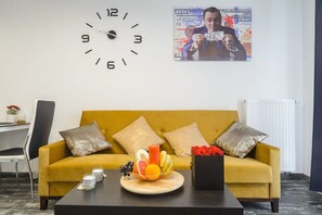Ein Zimmer mit einem gelben Sofa, einer Uhr, einem Poster, einer Obstschale und einem schwarzen Couchtisch.