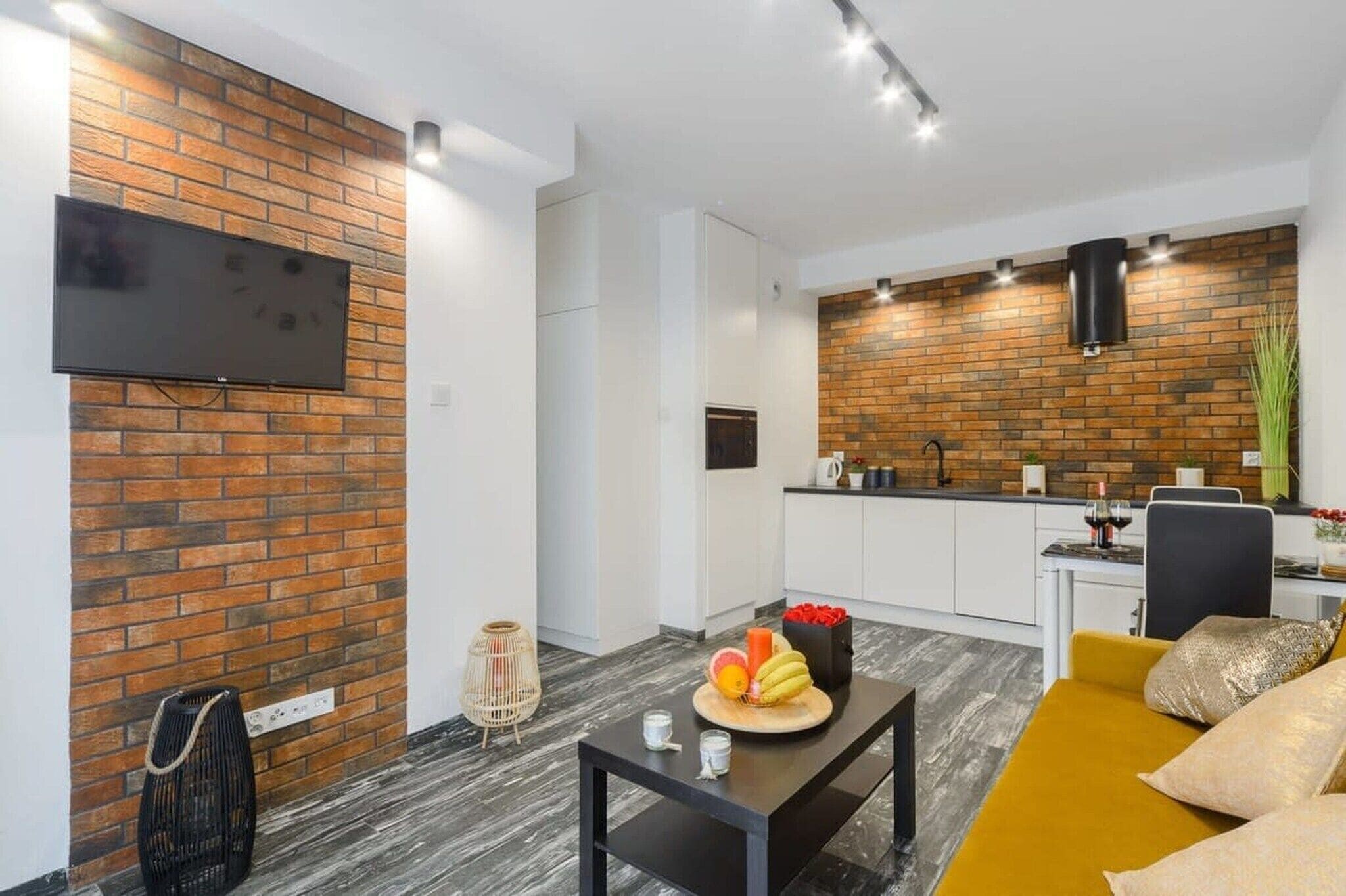 A modern living room with a brick accent wall, a flat-screen TV mounted above it, a dining area with a table set for two, and a kitchen with a brick backsplash and modern appliances.