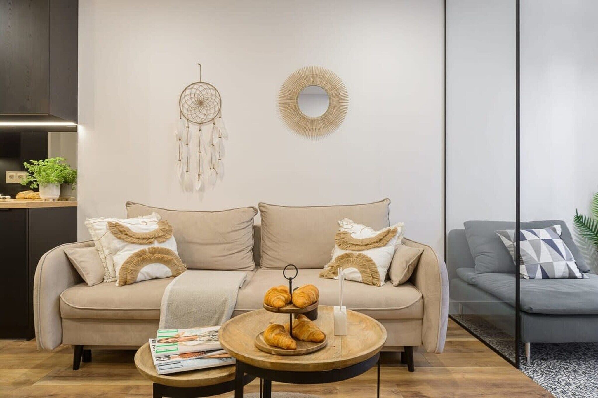 A modern living room with a beige sofa, two pillows, a round coffee table with a croissant holder, a dreamcatcher, and a mirror on the wall.