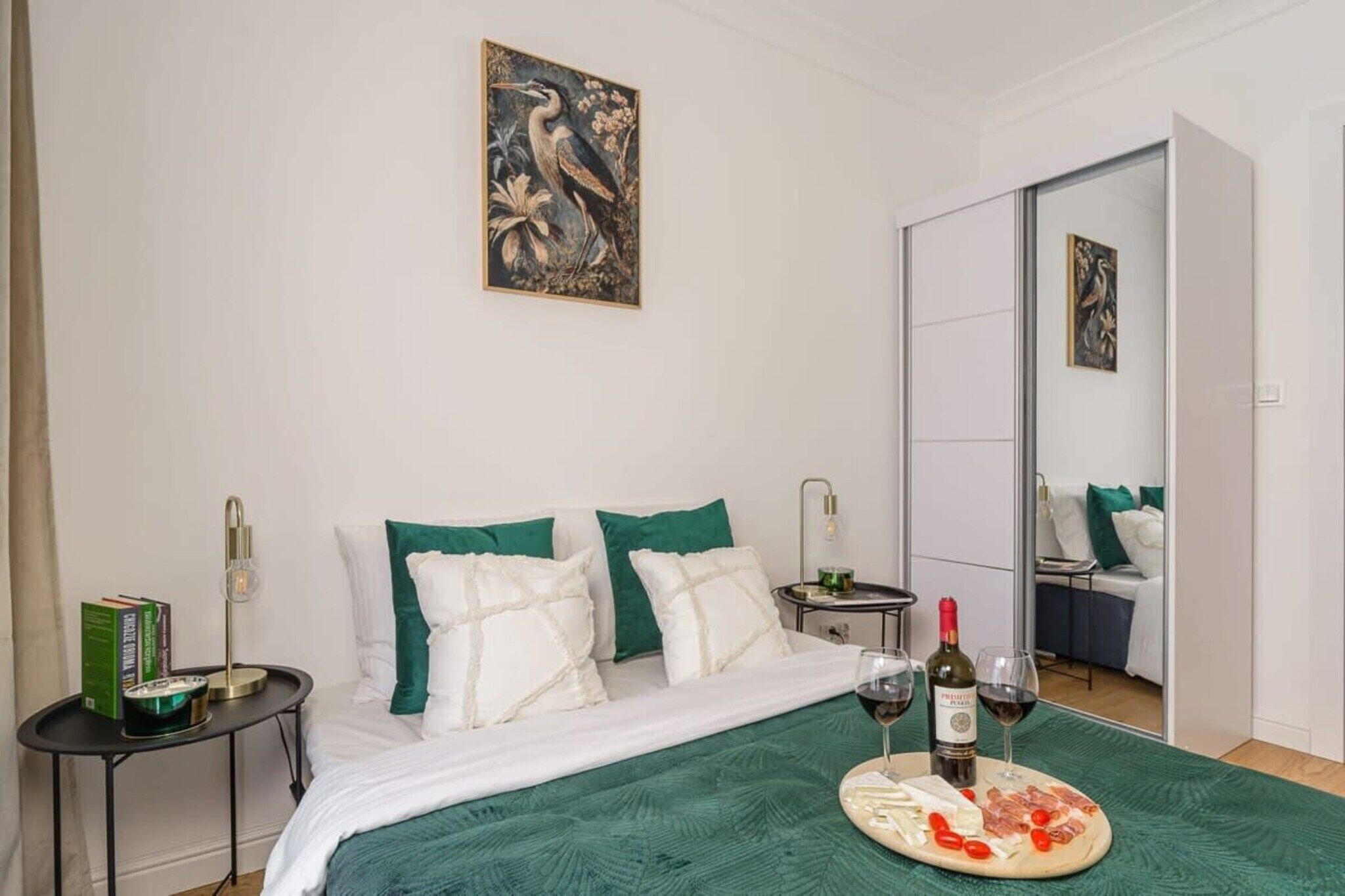 A neatly arranged bedroom with a bed, pillows, a bedside table with a lamp, a small table with a bottle and glasses, and a painting on the wall.